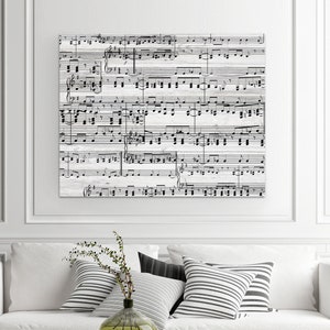 May include: A square canvas print featuring a detailed musical score in black against a white, weathered wood-like background. The artwork is mounted on a white wall above a white sofa with striped pillows and a black coffee table.