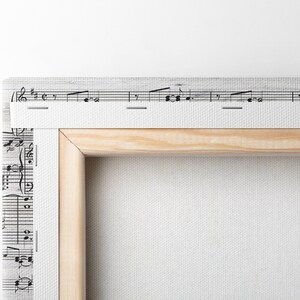 May include: A canvas art print featuring musical notes and a wooden frame. The print has a white background with black musical notation. The wooden frame is light brown. A metal screw is visible on the side.