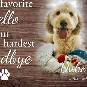 Op de afbeelding: Een lichte hond met zijn tong uit, rustend op een deken met een speeltje. De afbeelding bevat de tekst "hello", "hardest goodbye", en de naam "Duke" met de jaren "2008-2020". De achtergrond is een houten textuur.