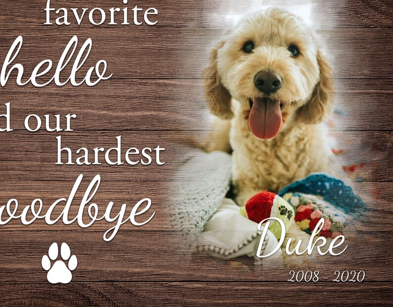 Op de afbeelding: Een lichte hond met zijn tong uit, rustend op een deken met een speeltje. De afbeelding bevat de tekst "hello", "hardest goodbye", en de naam "Duke" met de jaren "2008-2020". De achtergrond is een houten textuur.
