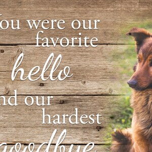 Op de afbeelding: Een houten bord met de tekst "Je was onze favoriete hallo en ons moeilijkste afscheid." Een bruin-zwarte hond staat aan de rechterkant van het bord. De achtergrond is een houten textuur.