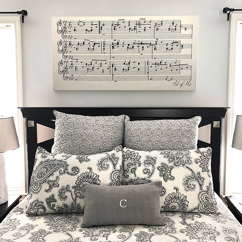 May include: A black bed frame with a white and gray patterned duvet and pillows. The headboard has a decorative molding. A framed print of sheet music with the words "All of Me" is hanging above the bed.