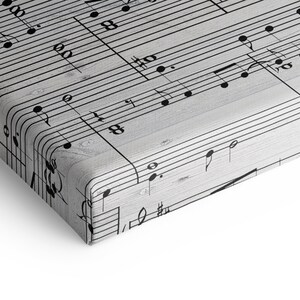 May include: A canvas print featuring a close-up of musical notes and symbols. The black notes are set against a light gray, wood-grain background. The print has rounded corners and a white border.