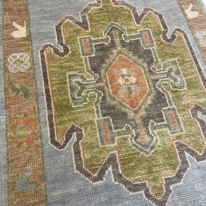 May include: A close-up of a blue rug with an intricate geometric pattern. The rug features a central design in shades of green, brown, and orange, outlined in dark gray. The border has a brown and tan design. The rug has a textured appearance.