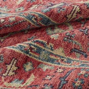 May include: Close-up of a red rug with a detailed pattern. The rug features a repeating design in shades of red, blue, and beige. The texture appears soft and plush, with visible woven details. The rug is folded, showing the depth of the material.