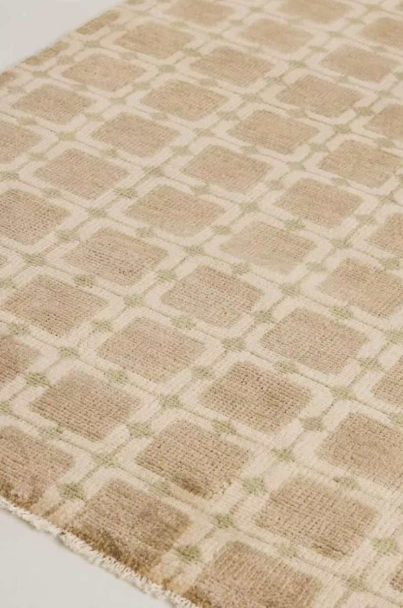 May include: Close-up of a beige rug with a repeating geometric pattern. The design features squares within squares, outlined in a lighter beige and pale green. The rug has a textured, woven appearance.