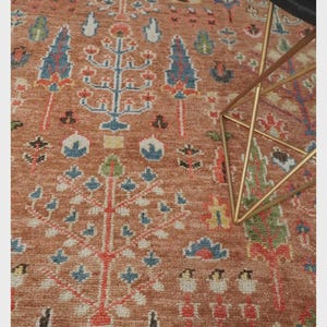 May include: A detailed close-up of a terracotta-colored rug with an intricate tree of life design. The rug features blue, red, green, and white accents. A gold-colored metal table leg and black tabletop are in the corner.