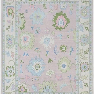May include: A rectangular area rug with a soft pink background and a floral pattern. The rug features a border with a repeating design and is adorned with various pastel-colored floral motifs, including blue, green, and pink. The rug has a fringed edge.
