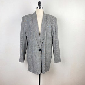 Può includere: Un blazer grigio con un sottile motivo a quadri. Il blazer ha una chiusura con un solo bottone e un colletto lungo e appuntito.