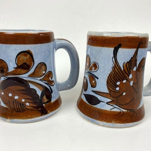 May include: Two ceramic mugs with a light blue base and brown rim and handle. Each mug features a unique brown and black bird and floral design. The mugs are hand-painted and have a rustic, artisanal feel.
