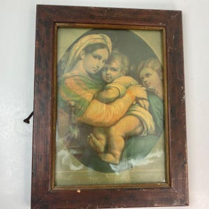 May include: A framed print of the Holy Family. The image features a woman holding a child, with another figure looking on. The print has a soft color palette of greens, yellows, and browns. The frame is made of dark wood.
