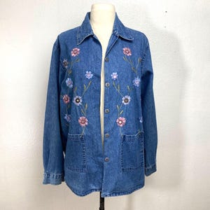 Può includere: Una giacca di jeans blu con fiori ricamati in tonalità rosa, blu e bianco. I fiori sono disposti in un motivo verticale sul davanti della giacca.