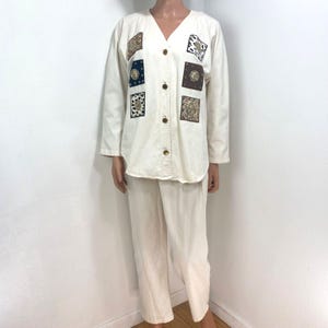 May include: A cream-colored two-piece set consisting of a long-sleeved button-down shirt and matching trousers. The shirt features a V-neckline and six square patches with varied animal print and metallic patterns arranged on the front panels.