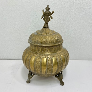 May include: A vintage brass container with a lid, featuring intricate embossed patterns and a small figure on top. The container rests on three ornate feet. The overall color is a warm, aged gold.