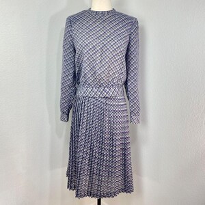 May include: A vintage dress with a long-sleeved top and pleated skirt. The dress features a checkered pattern in shades of purple, gray, and white. The top has a round neckline and the skirt falls to the mid-calf.