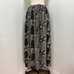 Puede incluir: Una falda larga hasta el suelo con un diseño de patchwork que presenta paneles alternos de patrones en blanco y negro, incluyendo motivos florales, lunares y rayas diagonales. La prenda tiene una cintura elástica y una silueta amplia y fluida.
