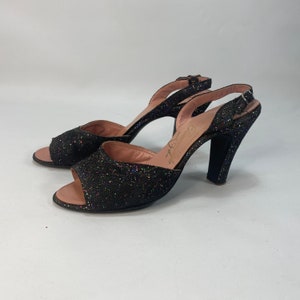 May include: A pair of black sparkly heeled sandals with a peep toe and an ankle strap. The sandals have a high heel and are made of a sparkly fabric.