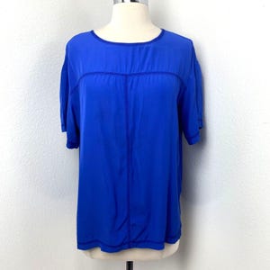 Puede incluir: Una camiseta azul brillante de manga corta. Tiene un cuello redondo y una costura vertical central. La tela parece suave y ligera. La camiseta se exhibe en un maniquí.