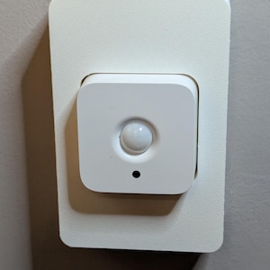 May include: A white motion sensor with a round lens and a small black dot in the centre. The sensor is mounted on a white square wall plate.