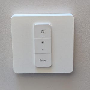 May include: A white wall-mounted smart light switch with a white faceplate and a rectangular button panel. The button panel has a power button, a brightness control, and a color temperature control. The button panel is labeled "hue".
