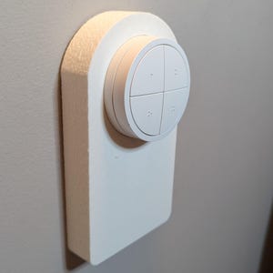 May include: White wall-mounted smart home device with four buttons. The device has a round, white face with four buttons arranged in a cross pattern. The device is mounted on a white wall.