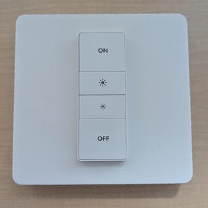 May include: A white smart light switch with four buttons. The top button reads "ON", followed by two light symbol buttons, and the bottom button reads "OFF". The switch is mounted on a square white plate.