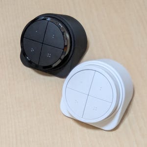 May include: Two smart home control devices. One is black with a silver trim, the other is white. Each has four black buttons and four small dots. The devices are circular with a square base.