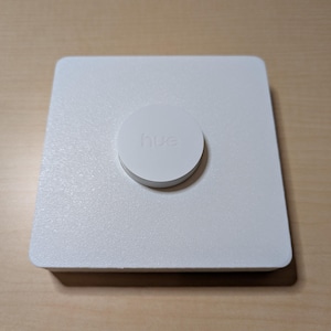 May include: A white square device with a circular button on top. The button is white and has the word "hue" printed on it. The device is sitting on a light brown wooden surface.