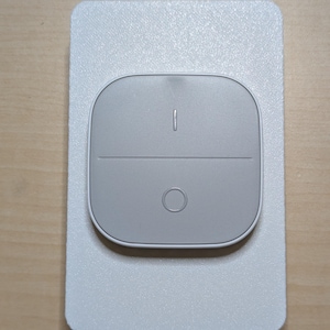 May include: A light gray smart home switch with a rectangular, textured faceplate. The switch has a rounded, gray control panel with a vertical line and a circle. The device is mounted on a wooden surface.