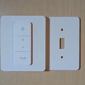 May include: A white smart light switch with three buttons and the brand name "hue" printed on it. The switch is mounted on a white square plate, with a separate white plate for a standard light switch.