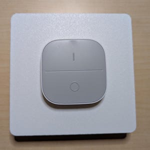 May include: A gray smart home control panel with a square white base. The panel has two buttons and a circular button. The top button has a vertical line. The panel is designed for home automation.