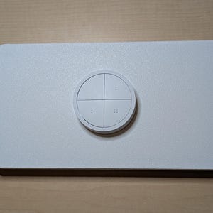 May include: A white rectangular control panel with a circular button in the center. The button is divided into four sections and has small raised dots. The panel has a textured surface.