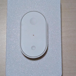 May include: A white rectangular device with a textured surface. It has a central, oval-shaped component with two circular indentations and a small red dot. The device is likely electronic.