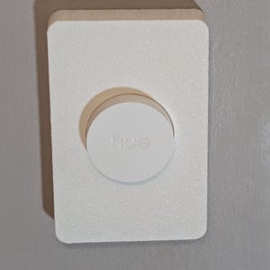 May include: A white, rectangular wall-mounted device with a circular button in the center. The button has the word "hue" printed on it. The device is mounted on a gray wall.