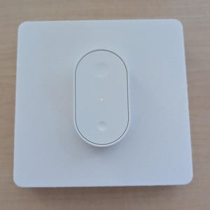 May include: A white smart home light switch, rectangular with a central oval button. The button has a small, yellow light illuminated. The switch is mounted on a light-coloured surface.