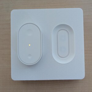 May include: A white, rectangular smart home device with two oval-shaped sections. One section has a button with a small yellow light, and the other is a recessed area. The device is designed for home automation.