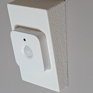 May include: A white motion sensor light with the word "hue" printed on it, mounted on a white wall.