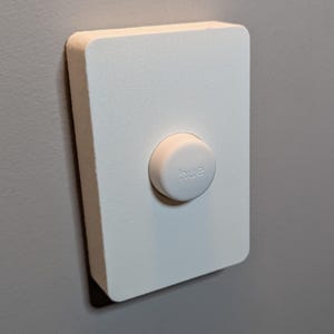 Peut inclure: Un appareil domotique rectangulaire blanc avec un bouton circulaire. Le bouton porte l'inscription "hue". L'appareil est monté sur un mur gris.