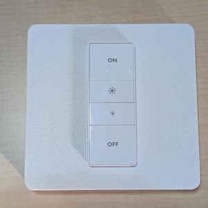 May include: A white smart light switch with four buttons. The top button reads "ON", followed by two buttons with a sun symbol, and the bottom button reads "OFF". The switch is mounted on a square, textured white plate.