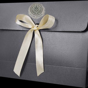 Op de afbeelding: Een donkergrijze cadeauzak met een crèmekleurig satijnen lint dat in een strik is geknoopt. De tas heeft een klepsluiting en een cirkelvormig logo met de tekst "LUXE VERDE". De tas is gemaakt van een textuurrijk materiaal.