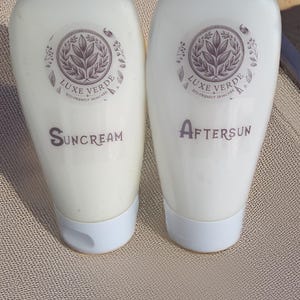 Puede incluir: Dos tubos blancos de productos para el cuidado de la piel ecológicos Luxe Verde, uno con la etiqueta "Suncream" y el otro "Aftersun". Cada tubo tiene una tapa blanca y un logotipo circular con un diseño de hojas. Los productos están sobre una superficie beige.