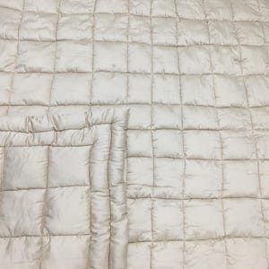 Può includere: Una coperta trapuntata color crema con un motivo a rombi. La coperta è piegata a metà, mostrando il dettaglio delle cuciture.