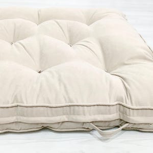 Op de afbeelding: Een beige, gecapitonneerde futonmatras met een rechthoekige vorm. De matras heeft een gewatteerd ontwerp met meerdere knoopaccenten. De randen zijn afgewerkt met een contrasterende bruine bies en een stoffen lus.