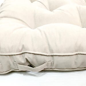 Könnte beinhalten: Ein beige getuftetes Kissen mit Griff. Das Kissen hat eine rechteckige Form mit abgerundeten Ecken und einem abgesteppten Rand. Der Stoff ist eine weiche, neutrale Farbe, geeignet für den Innen- oder Außenbereich.