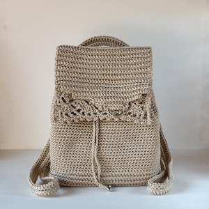 Può includere: Uno zaino beige all'uncinetto con chiusura a patta e coulisse. La borsa presenta un design decorativo all'uncinetto sulla patta e spallacci regolabili. Lo zaino ha una maniglia superiore.