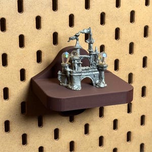 May include: A miniature silver castle figurine with two crystal-like orbs, displayed on a small, dark brown shelf. The shelf is mounted on a pegboard with evenly spaced oval holes. The castle has intricate architectural details.