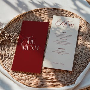 以下が含まれることがあります： 織られたトレイに2つのメニューカード。1枚は白の筆記体で「THE MENU」と書かれたバーガンディ色。もう1枚はオフホワイトで、バーガンディ色の筆記体で「MENU」とメニュー項目が記載されています。メニューには、ラム、サーモン、アボカドスープ、ティラミスが含まれています。