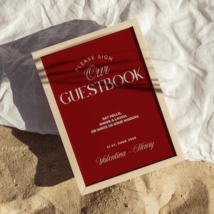Puede incluir: Un letrero de libro de visitas enmarcado con un fondo rojo y un marco color crema. El letrero dice "PLEASE SIGN Our GUESTBOOK" en una escritura elegante. También dice "SAY HELLO, SHARE A LAUGH, OR WRITE US SOME WISDOM." La fecha es el 21 de junio de 2026, con los nombres Valentina y Henry.