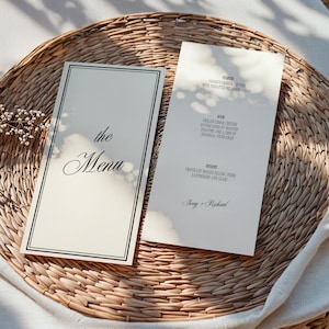 Puede incluir: Dos tarjetas de men&uacute; color marfil con texto negro y borde negro, sobre una bandeja de mimbre tejido. Una tarjeta dice "the Menu" en escritura cursiva. La otra enumera un entrante, un plato principal y un postre, con "Ivory + Richard" abajo.