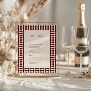 Pode incluir: Uma placa de vidro transparente com uma borda xadrez bordô e branca exibe "the Bar" e um menu de bebidas. Duas taças de vinho e uma garrafa de champanhe estão ao fundo. A placa lista bebidas exclusivas, vinho, cerveja e opções não alcoólicas.